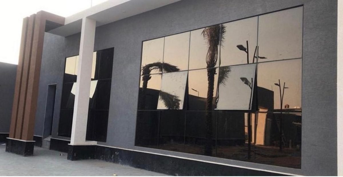 واجهة مبنى تجاري باللون الرمادي الداكن مركّب عليها ألواح زجاج عاكس كبيرة بإطارات، تنعكس على سطحها أشجار النخيل وأعمدة الإنارة وسماء الغروب.