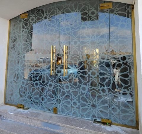 واجهة زجاجية مقوسة لمحل، مغطاة بنقش هندسي إسلامي مصنفر، وبها باب مزدوج من الزجاج بمفصلات ومقابض نحاسية اللون